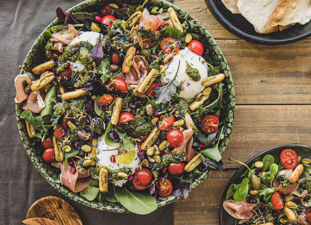 Kreativer salat mit Parma Schinken, Burrata und Grissini Sticks. Auf dem Salat hat es eine grüne Pestosauce und Olivenöl Auf einem Tisch dazu Besteck aus Olivenholz.